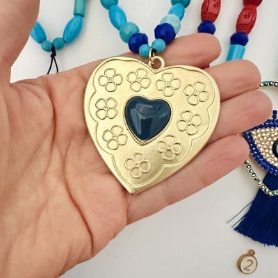 Colar com pendente em forma de coração dourado decorado com flores e coração azul no centro, segurado numa mão.