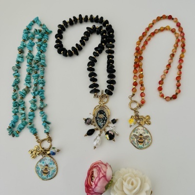 Três colares com contas coloridas e pendentes dourados decorativos, junto a duas flores decorativas