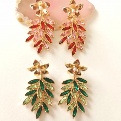 Duas pares de brincos folhas douradas com pedras vermelhas e verdes, com flores douradas e pérolas, sobre fundo branco e concha rosa.