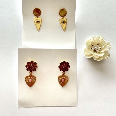Dois pares de brincos dourados com detalhes pretos, vermelhos e rosa em cartões brancos, junto a flor decorativa branca com pérolas pequenas.
