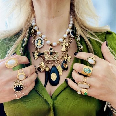 Colar com contas de pérola e pendentes dourados e pretos, anéis dourados com pedras, vestido verde com textura.