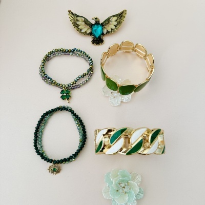 Cinco peças de bijuteria com detalhes verdes, brancos e dourados sobre fundo claro.
