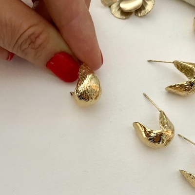 Brincos dourados martelados em forma de gota sendo segurados por mão com unhas vermelhas