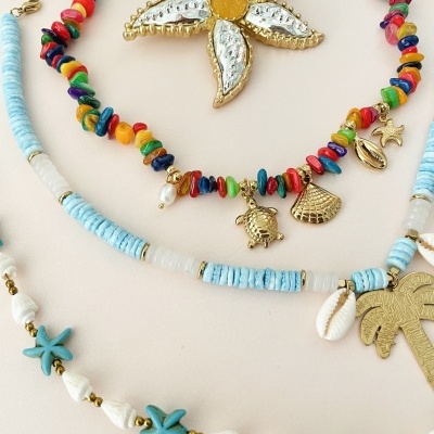 Três colares coloridos com contas e pendentes dourados sobre fundo neutro