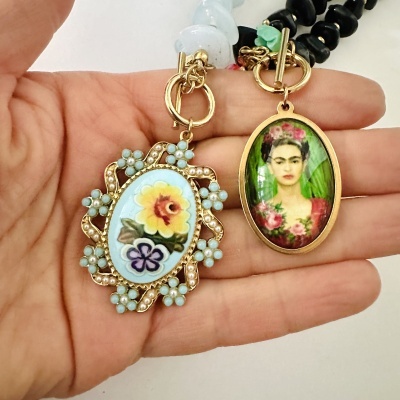 Dois pendentes ovais emoldurados em dourado com imagens decorativas, sobre uma mão aberta.