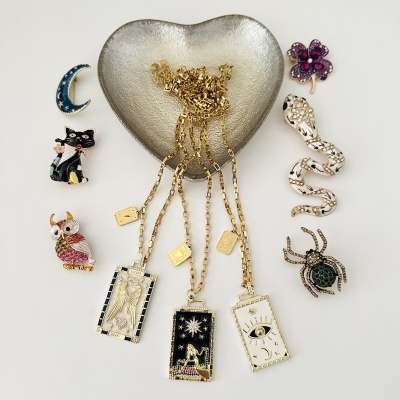 Colares dourados com pendentes retangulares com esmalte e cristais em prato em forma de coração prateado, rodeados por pins decorativos coloridos e com cristais.