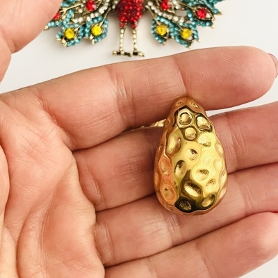 Peça dourada com padrão amolgadela na mão e broche pavão colorido ao fundo