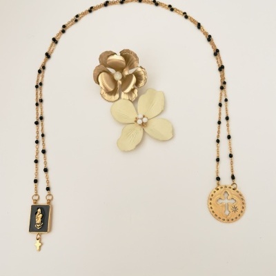 Colar terço dourado com contas pretas e pendentes religiosos, sobre fundo branco com duas flores decorativas.