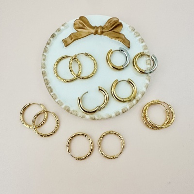 Brincos de argola dourados e prateados com textura, sobre base branca com laço dourado