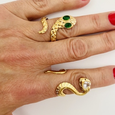 Mão com três anéis dourados em forma de serpente, dois têm olhos verdes e um tem uma pedra branca, unhas vermelhas.
