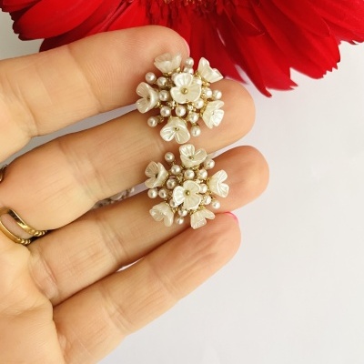 Brincos florais cor de pérola segurados pela mão com flor vermelha ao fundo