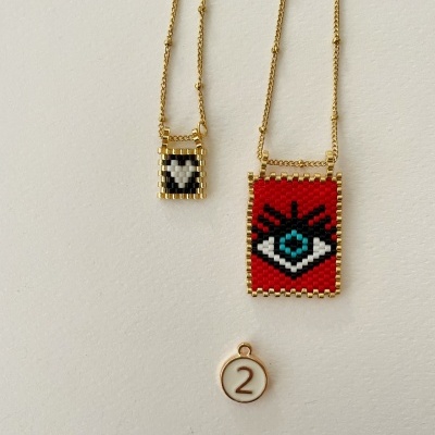 Dois colares dourados com pendentes quadrados em padrão mosaico, um vermelho com olho e outro xadrez, ambos pendurados numa superfície clara, com uma medalha branca e dourada com número 2