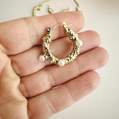 Brinco dourado em forma de argola com pérolas e textura irregular segurado por uma mão
