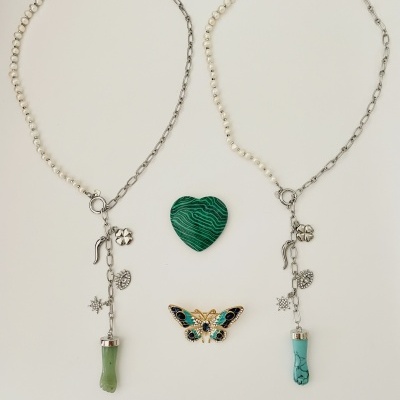 Dois colares com correntes e pérolas, um coração verde com linhas e um broche de borboleta colorido sobre fundo branco