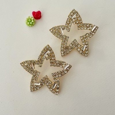 Brincos em formato de estrela com brilhantes, sobre fundo branco, com duas pequenas flores decorativas ao lado.