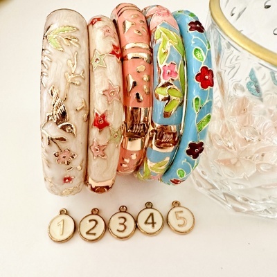 Conjunto de cinco pulseiras esmaltadas coloridas com padrões florais e medalhas numeradas ao lado de copo de vidro
