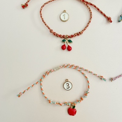 Duas pulseiras trançadas com pendentes de frutas, cerejas e maçã, em fundo branco.