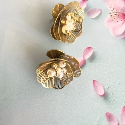 Brincos dourados com pérolas em forma de flor, sobre fundo azul claro com pétalas cor-de-rosa