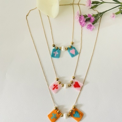 Três colares dourados com pendentes de contas coloridas e flores ao fundo