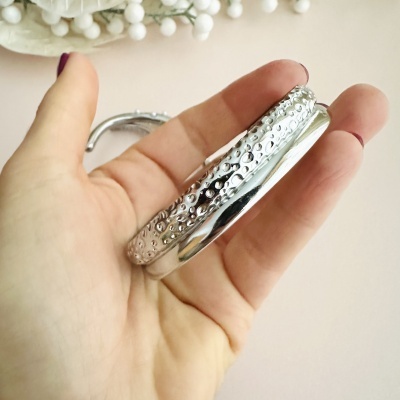 Pulseira prateada lisa e texturizada na mão, com unhas roxas, fundo rosa