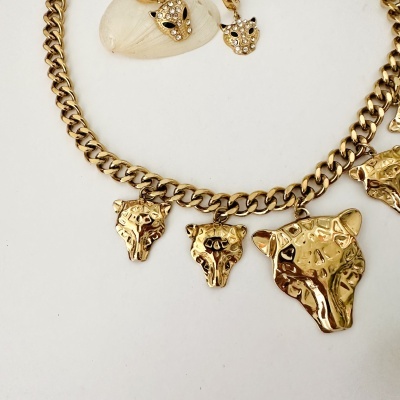 Colar e brincos dourados com cabeças de leopardo e pedras, sobre fundo branco com concha.