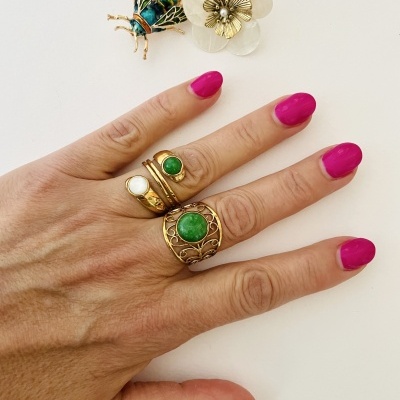 Mão feminina com três anéis em tons dourados e pedras verdes e branca, unhas pintadas rosa, sobre mesa branca com broches abelha e flor.