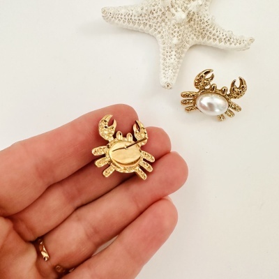 Broches dourados em forma de caranguejo, com uma pérola no centro, sobre fundo branco com estrela do mar