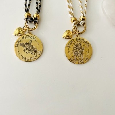 Duas pulseiras com medalhões dourados e cordões trançados, uma preto e branco outra branco e dourado