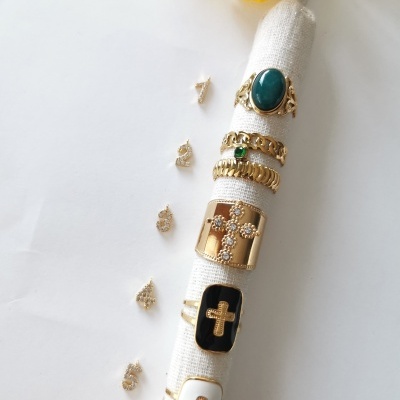 Anéis dourados variados com pedras e cruzes em suporte de tecido bege, fundo branco e flores amarelas