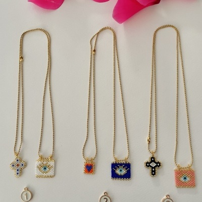 Colares dourados com pendentes coloridos de mosaicos em forma de cruz e retângulo, numerados 1 a 3, com flor rosa ao fundo