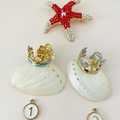 Duas coroas pequenas sobre conchas brancas, pendentes com números 1 e 2, estrela-do-mar vermelha com strass
