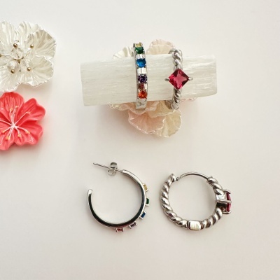 Anéis e brincos de prata com pedras coloridas e vermelhas em fundo claro com flores decorativas