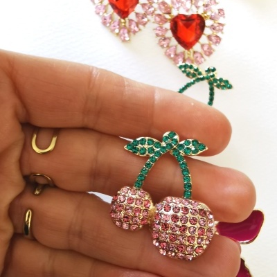 Brinco em forma de cerejas com pedras rosas e verdes, segurado por mão, fundo branco