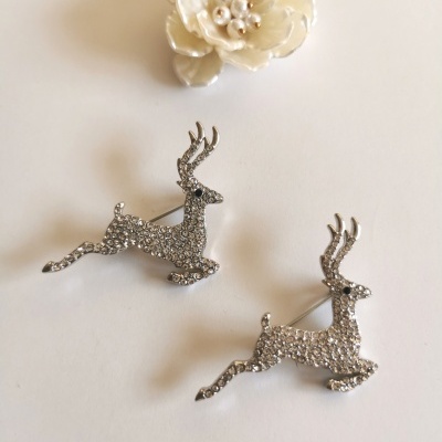 Dois broches em forma de veado prateado com pedras brilhantes e uma flor branca com pérolas sobre fundo branco