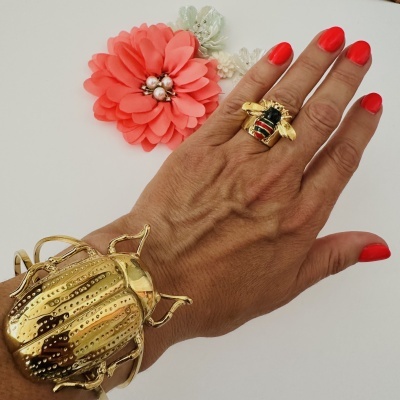 Mão com anel de abelha e bracelete escaravelho dourados e flores decorativas ao fundo