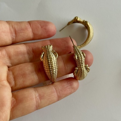 Brincos dourados com textura de crocodilo na mão sobre fundo branco