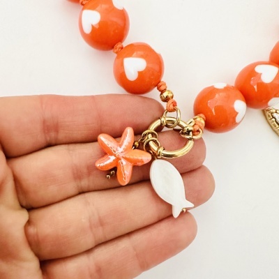 Colar laranja com corações brancos, pendentes estrela-do-mar e peixe, detalhes dourados, sobre mão