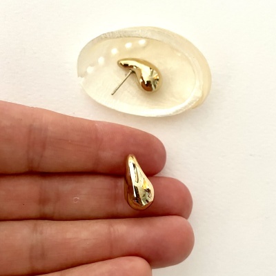 Par de brincos dourados em formato de gota, um sobre dedos e outro numa superfície branca translúcida.