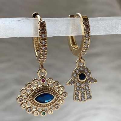 Brincos dourados com pedras, um em forma de olho azul e outro em forma de mão com cristais