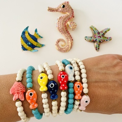 Pulseiras com contas coloridas e pendentes de peixe e mão, com três adereços marítimos brilhantes em fundo branco.