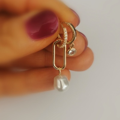 Brincos dourados com pérola e pedra transparente segurados por mão com unhas vinho