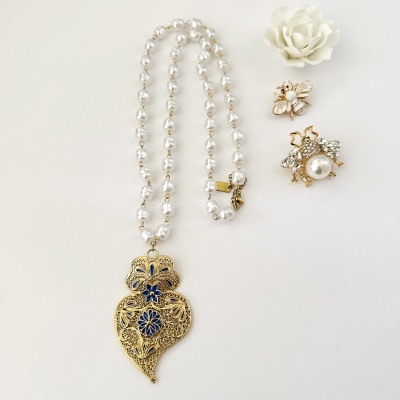 Colar de contas peroladas com pendente dourado florido azul e dois anéis dourados com pedras, sobre fundo branco com flor
