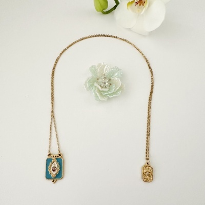 Colar dourado com dois pendentes retangulares e flores brancas ao fundo