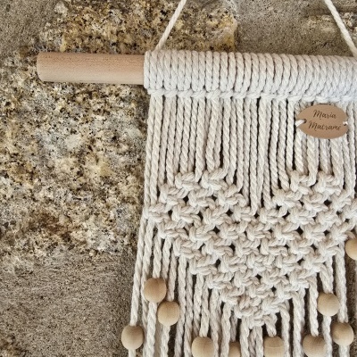 Tapeçaria de macramé branca com coração e contas de madeira pendurada numa parede de pedra
