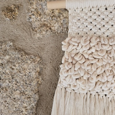 Parede com pedra aparente e pendurado decorativo de macramê cru com franjas longas.