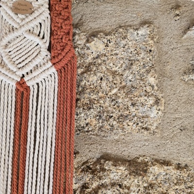 Tapeçaria macramé branca e castanho-avermelhado pendurada numa parede de pedra com etiqueta Mimi Maison.