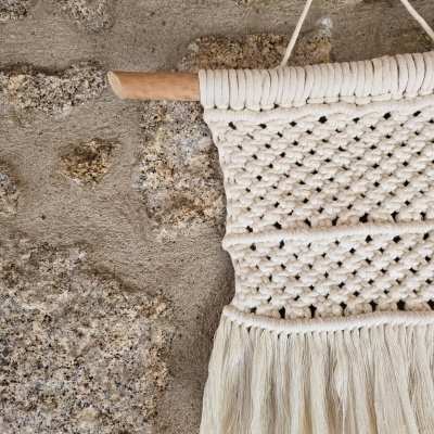 Tapeçaria em macramé creme com franjas pendurada numa parede de pedra