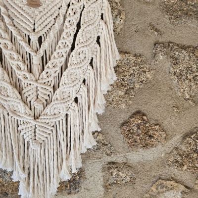Tapeçaria branca de macramé com desenhos em nós sobre parede de pedra