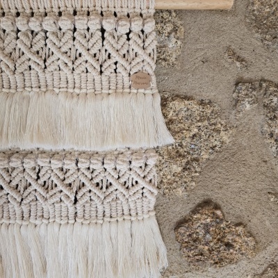 Tecido macramê creme com padrões e franjas pendurado numa parede de pedra