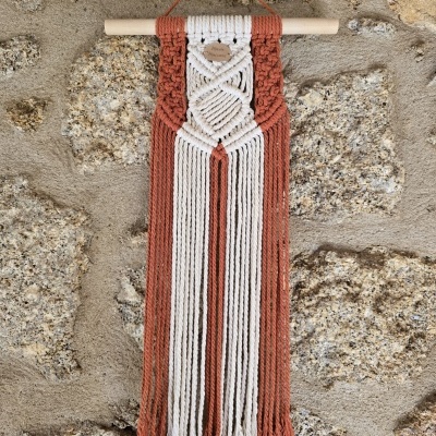 Pendurador de parede em macrame branco e terracota com franjas, pendurado numa parede de pedra.